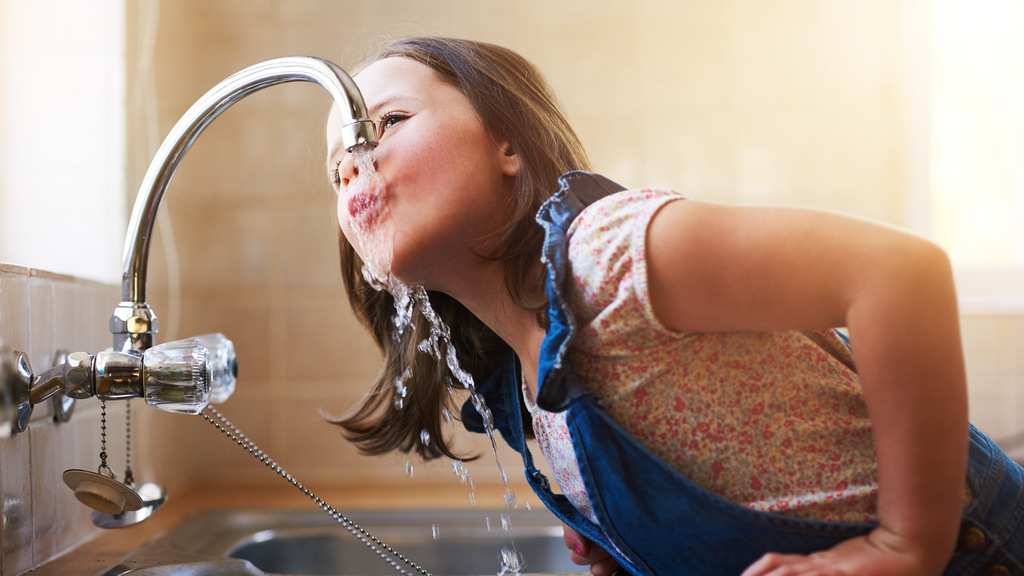 En flicka hänger över diskhon och dricker vatten direkt från vattenkranen.