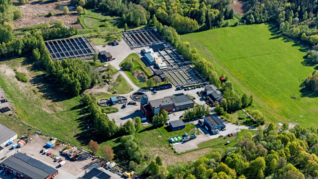 Flygbild över området där Gävles avloppsreningsverk ligger. Byggnaden för reningsverket samt reningsbassängerna omges av gröna fält och lövträd.